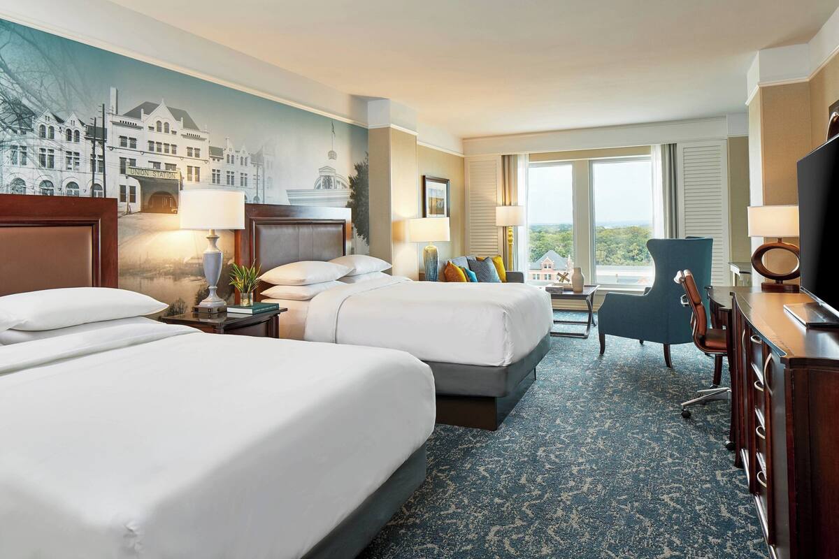 A hotel room with two beds, a desk, a chair, and a large window offering a city view.