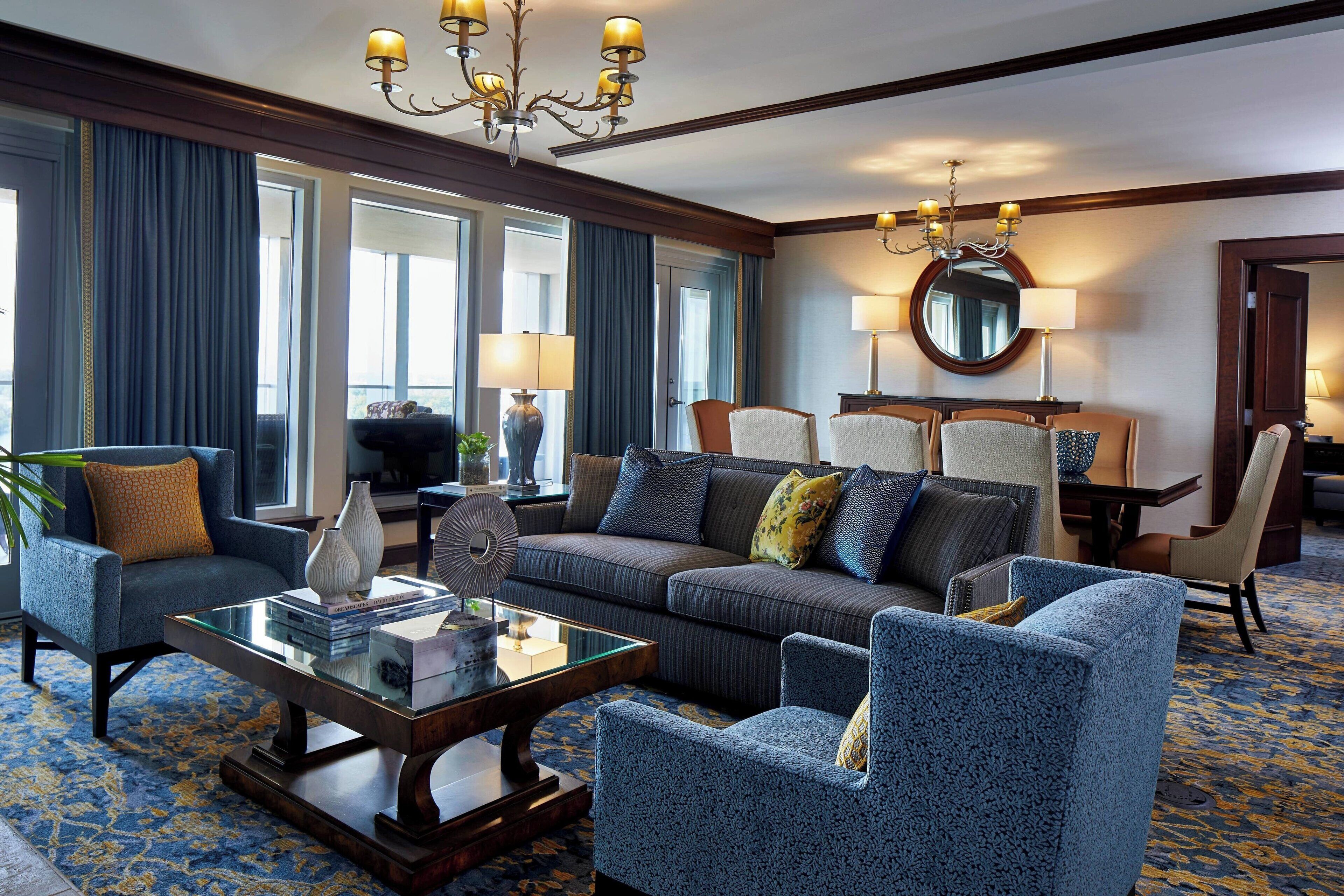 A hotel room with a large sofa, armchairs, a glass coffee table, and a dining area with chairs and a table.