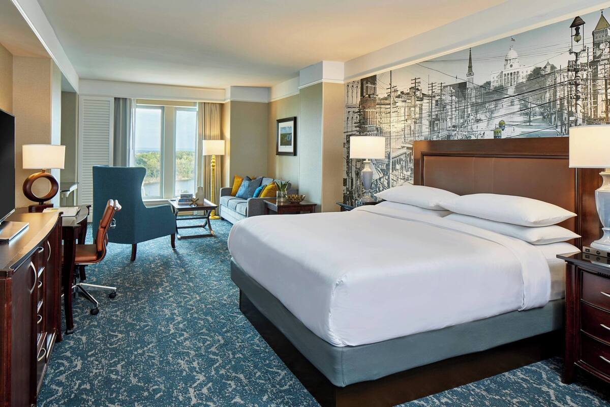 A hotel room with a large bed, a desk with a chair, a sofa, and a view of a cityscape through the window.