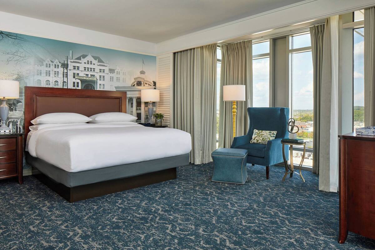 A hotel room with a large bed, a chair, a desk, and a view of the city through the windows.