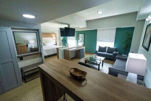 Una habitación de hotel moderna con kitchenette, zona de comedor, sala de estar con sofá y televisión, y un ventanal grande.