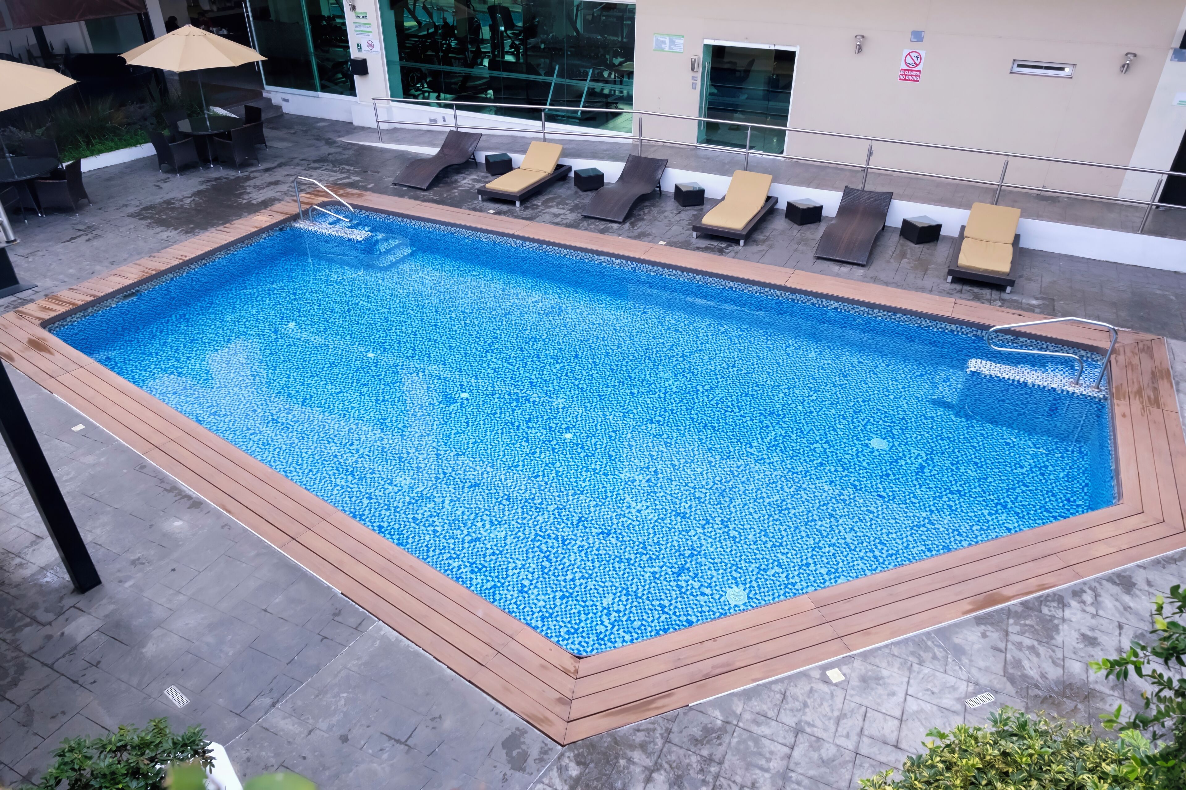 A rectangular pool with a wooden deck edge, surrounded by lounge chairs and a glass railing.