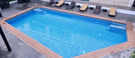 A rectangular pool with a wooden deck edge, surrounded by lounge chairs and a glass railing.