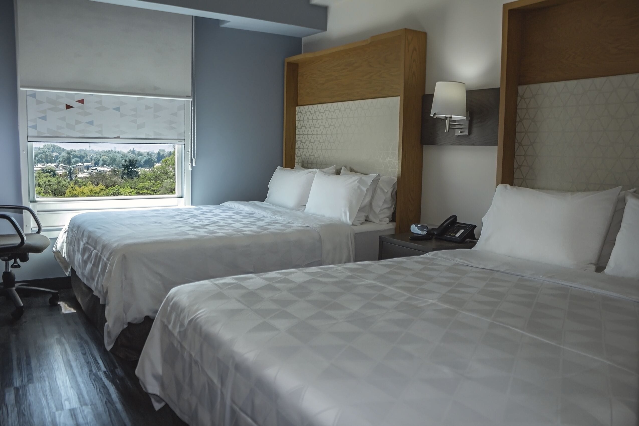 Ein Hotelzimmer mit zwei Betten, einem Schreibtisch, einem Stuhl und einem Fenster mit Blick auf die Stadt.