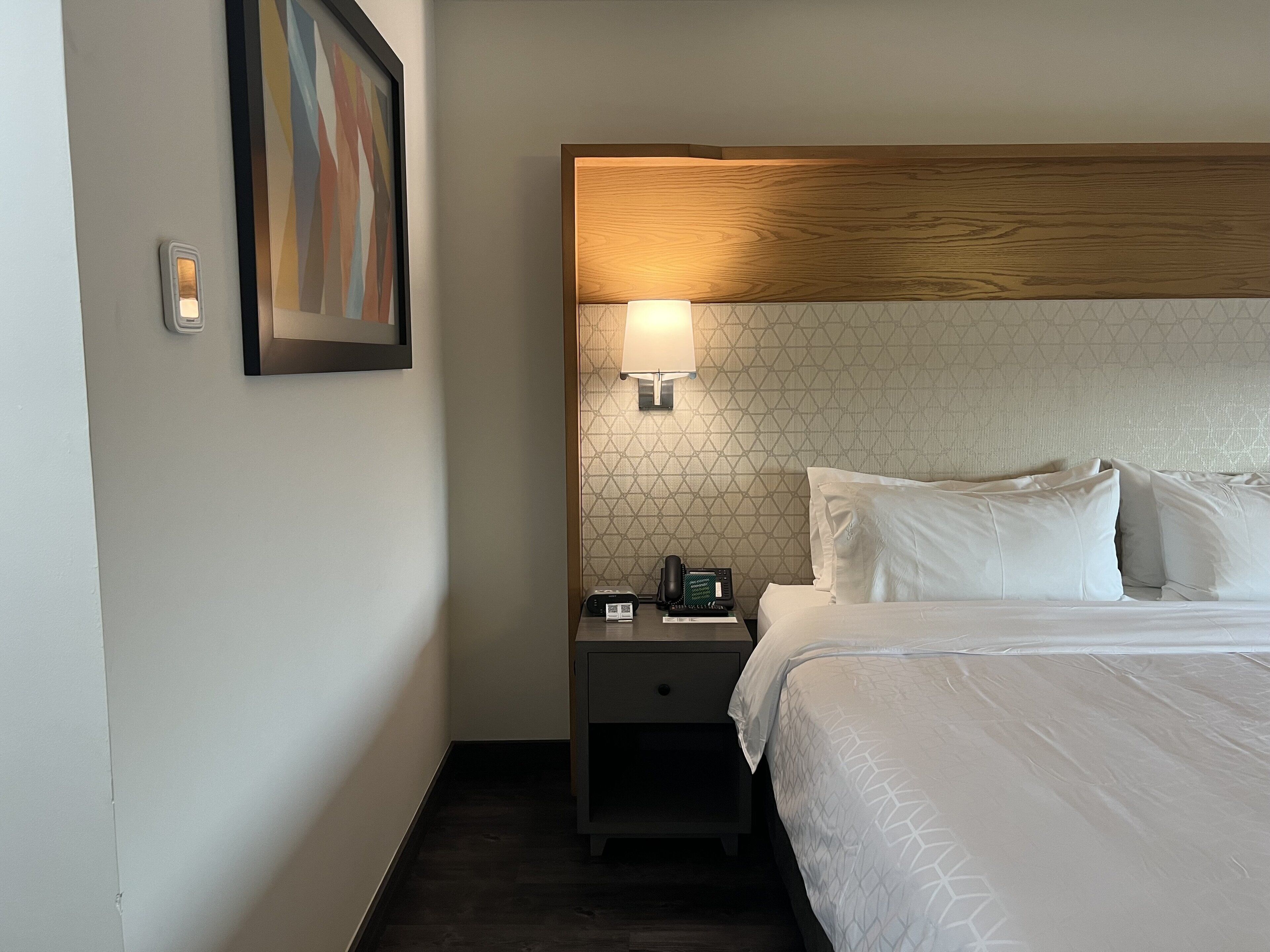 A hotel room with a neatly made bed, a bedside lamp, a telephone, and a framed picture on the wall.