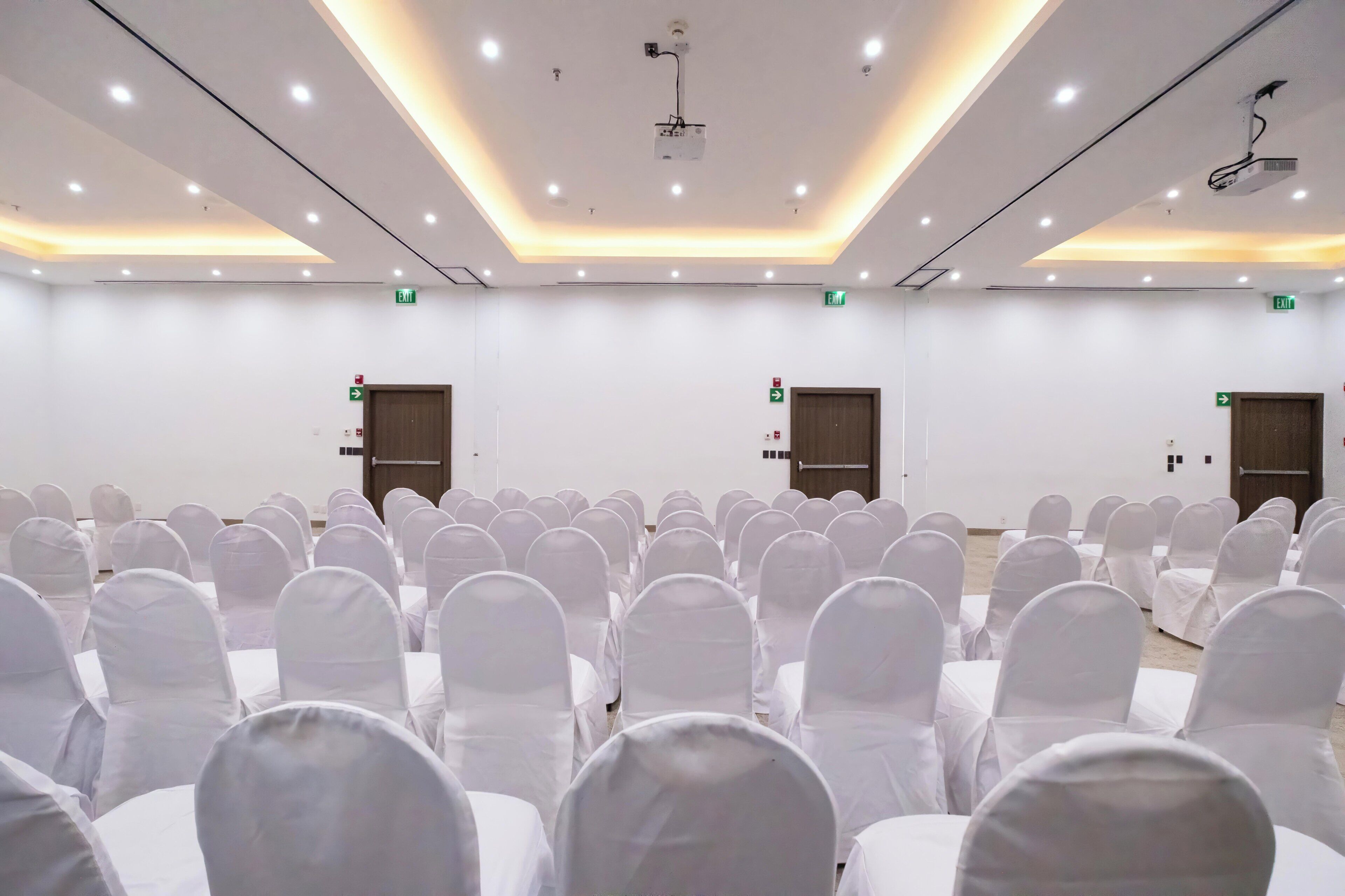 Un salón de conferencias con filas de sillas blancas, un proyector y luces en el techo.