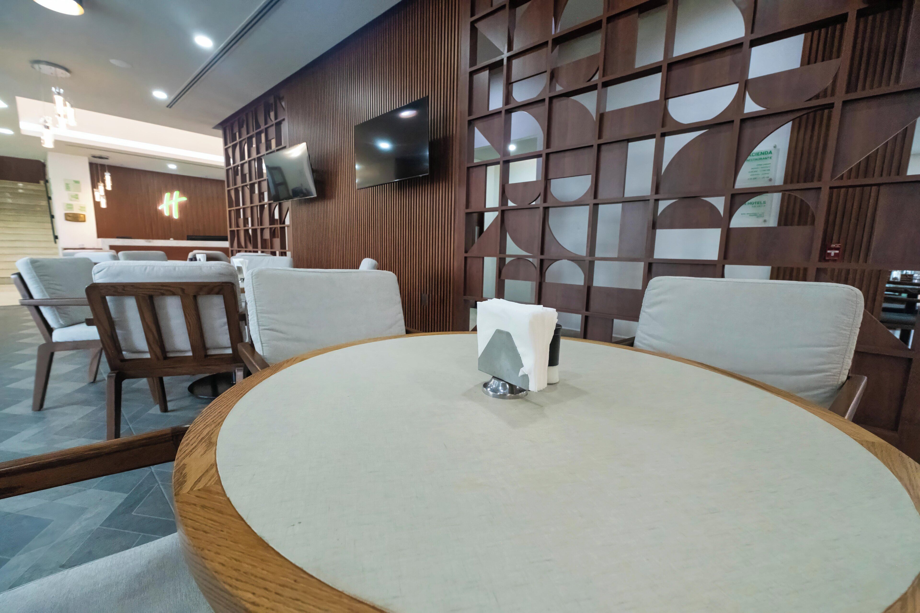 Un hall de hotel con una mesa redonda, sillas acolchadas, un televisor montado en la pared y un separador de madera con diseños geométricos.