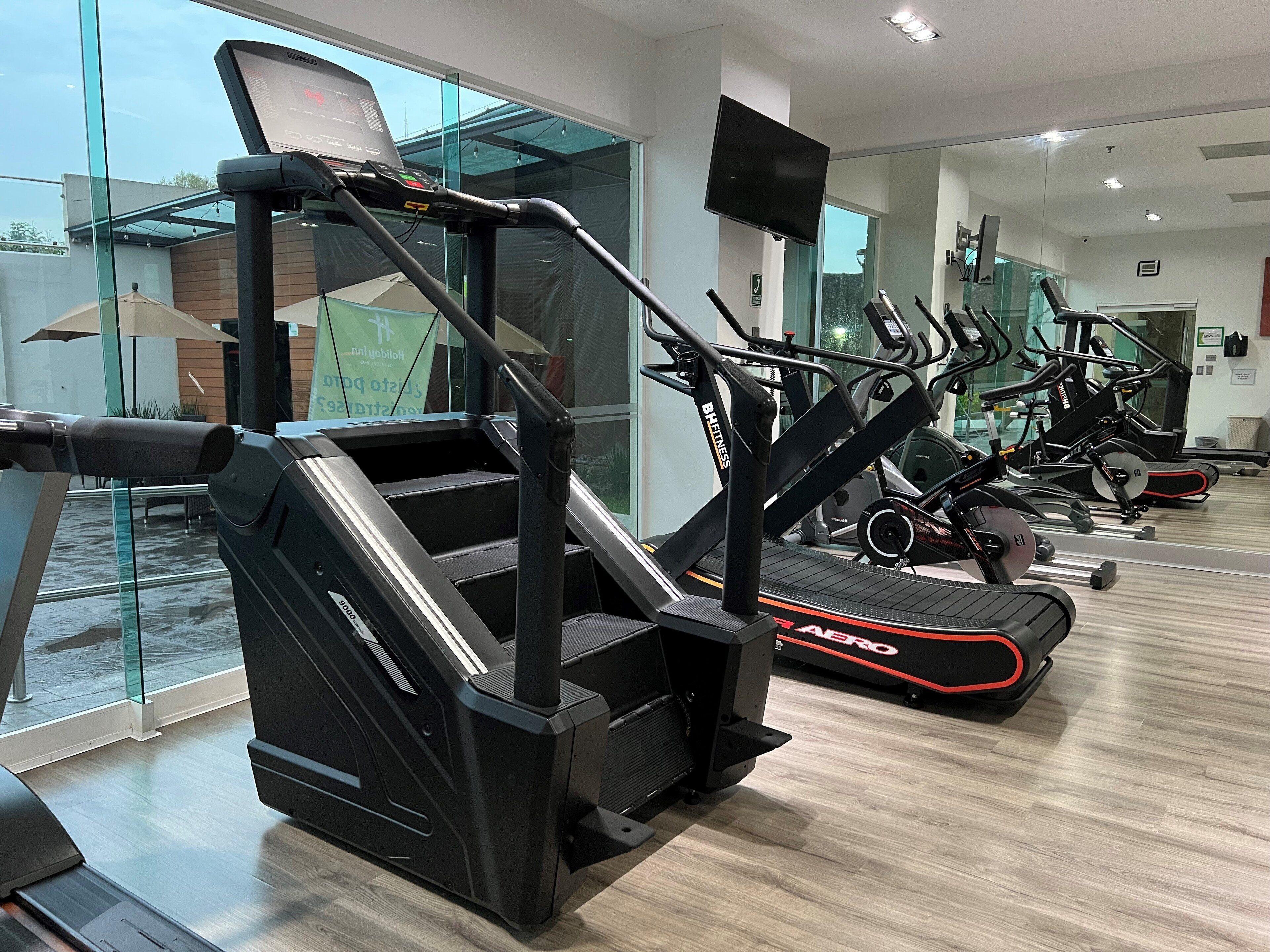 Ett gym med olika träningsmaskiner, inklusive löpband och en motionscykel, med ett stort fönster och en spegel som reflekterar utrustningen.