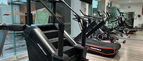 Ett gym med olika träningsmaskiner, inklusive löpband och en motionscykel, med ett stort fönster och en spegel som reflekterar utrustningen.