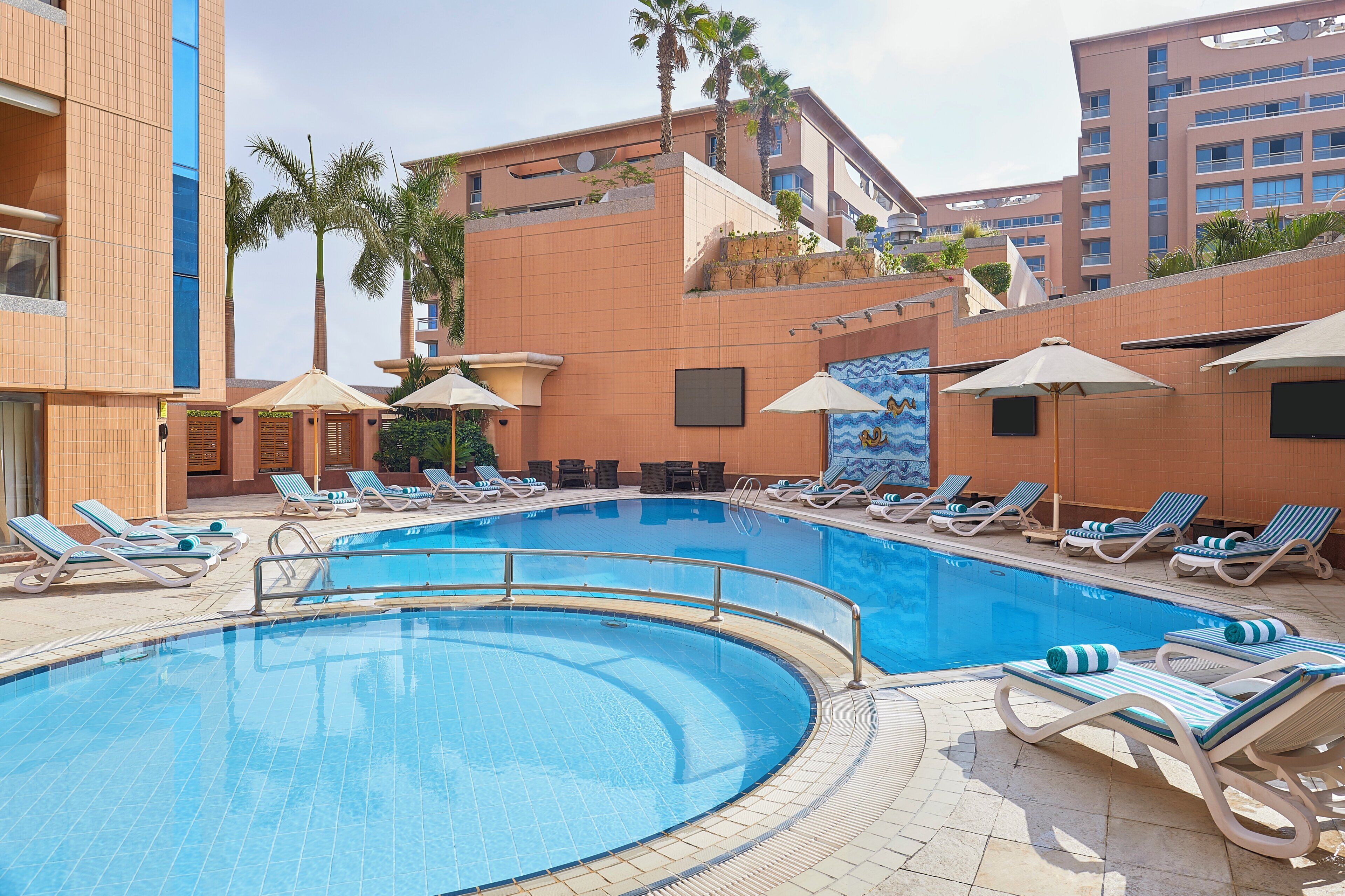 A pool area with lounge chairs and umbrellas, surrounded by buildings with balconies and palm trees.