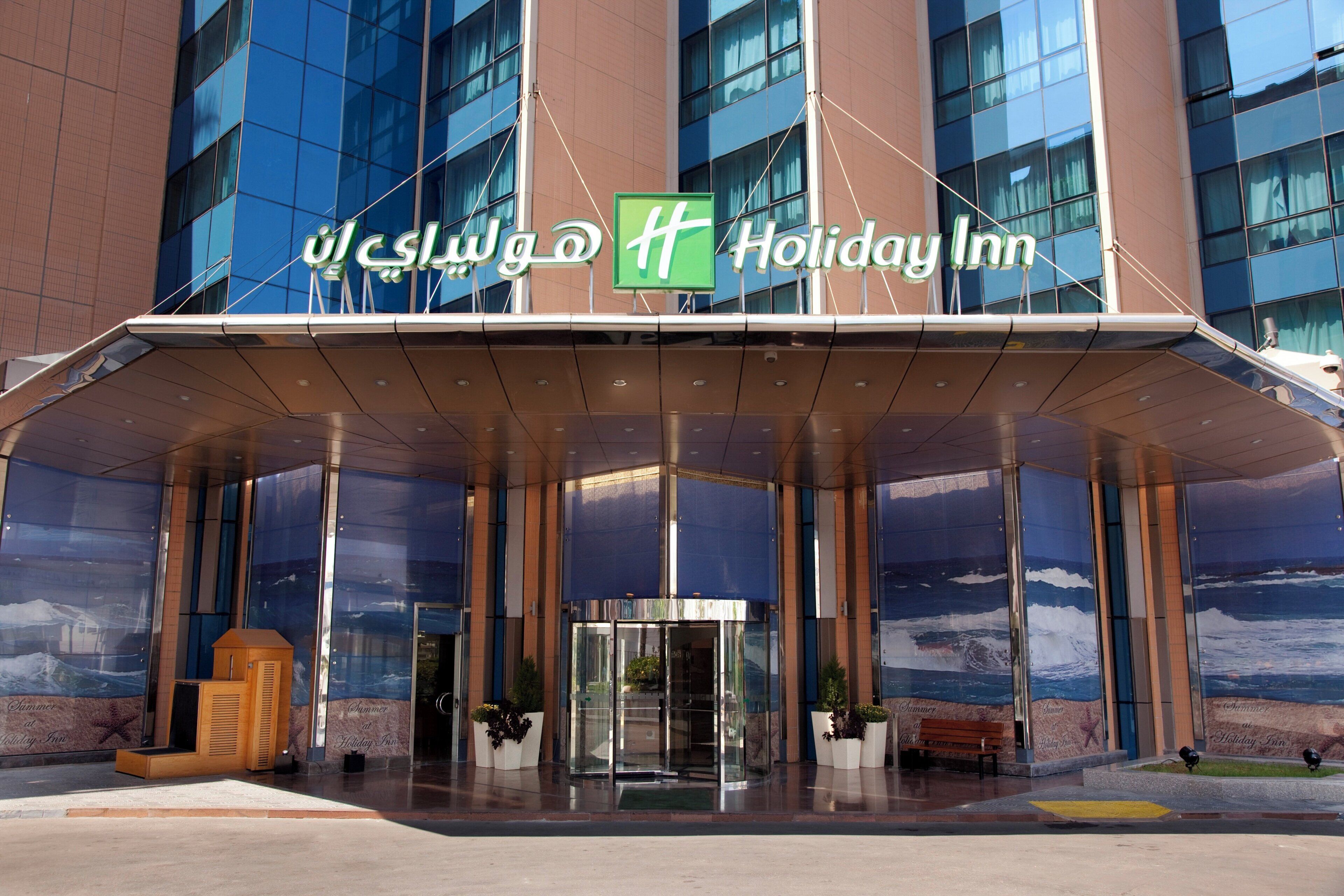 L’entrée d’un hôtel Holiday Inn, avec une façade en verre, un espace d’entrée abrité et une enseigne en anglais et en arabe.