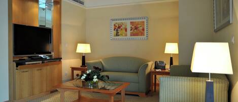 Una habitación de hotel con televisión, sofá, sillón, una mesa de centro con flores y dos lámparas.