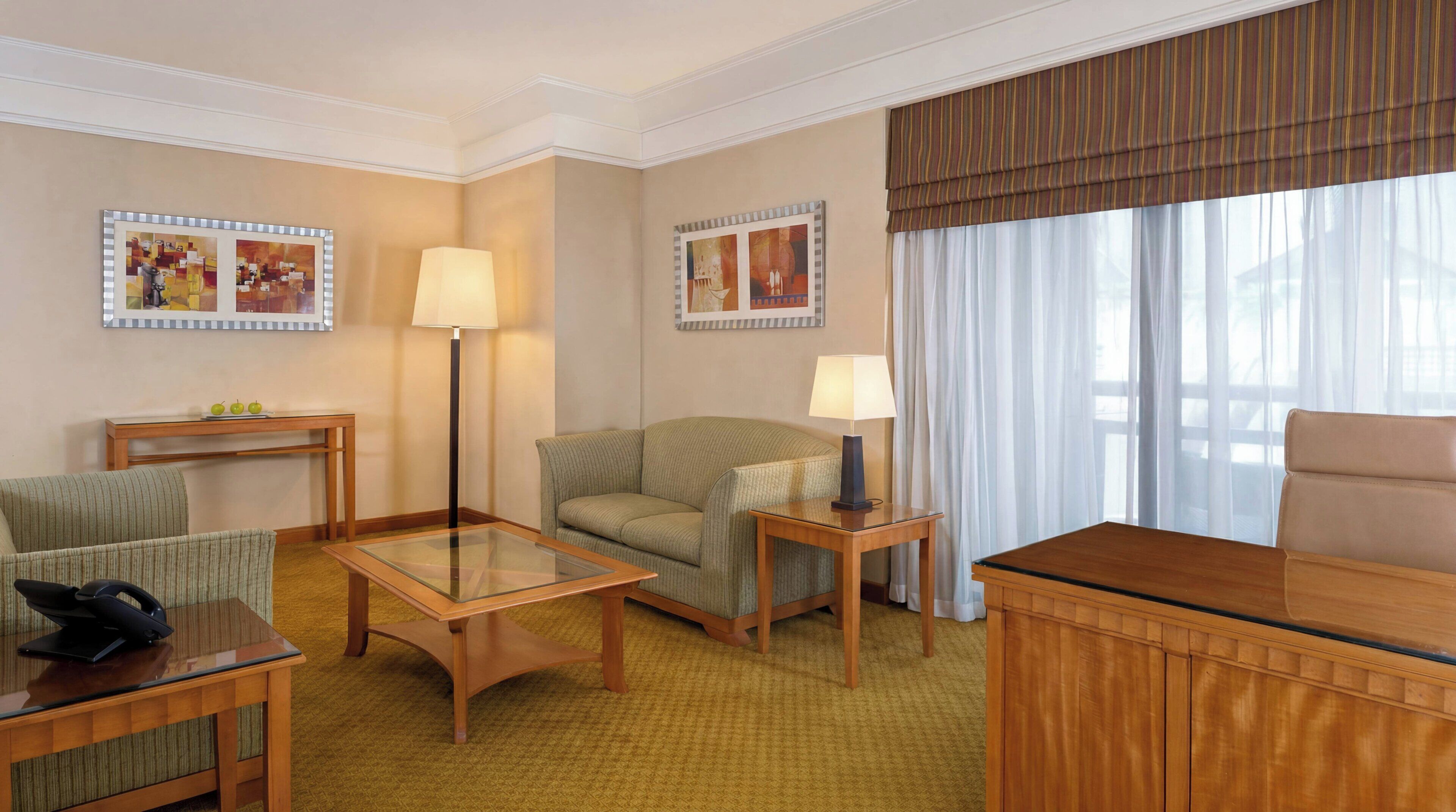 A hotel room with a sofa, armchair, glass-top table, lamp, and a desk with a telephone.