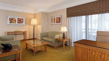 A hotel room with a sofa, armchair, glass-top table, lamp, and a desk with a telephone.