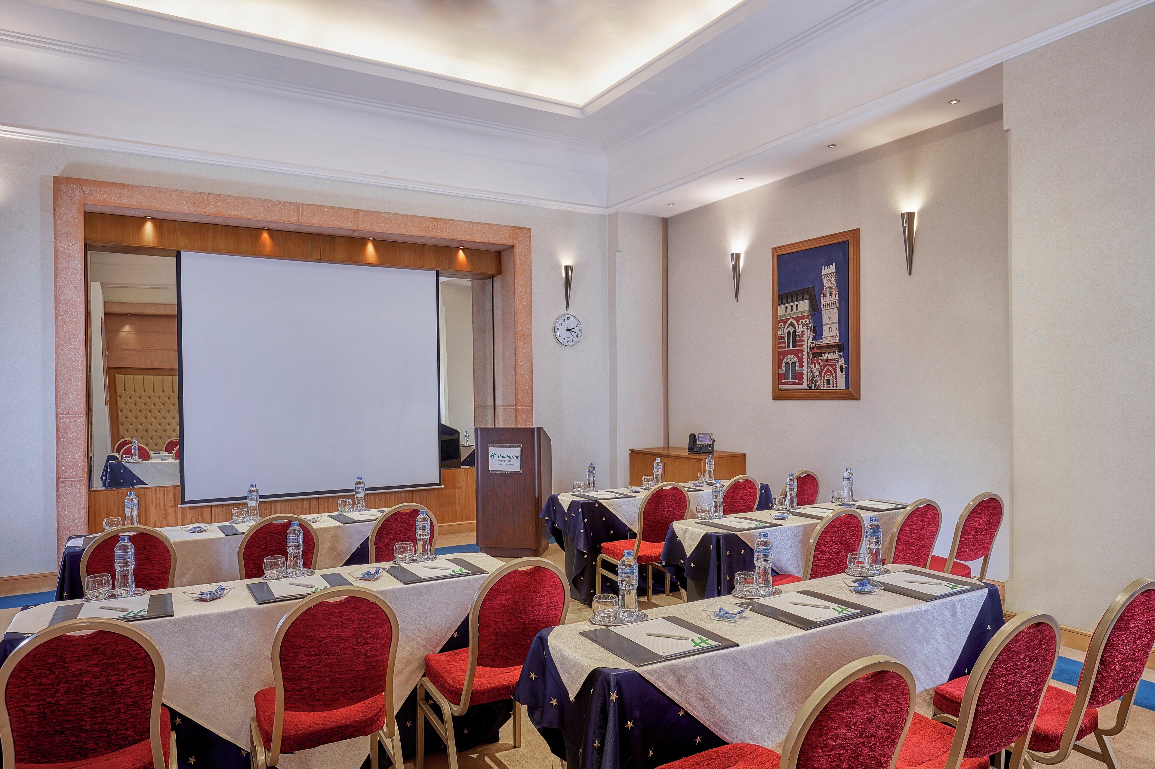 Une salle de conférence équipée de plusieurs tables, de chaises, d’un pupitre et d’un grand écran.