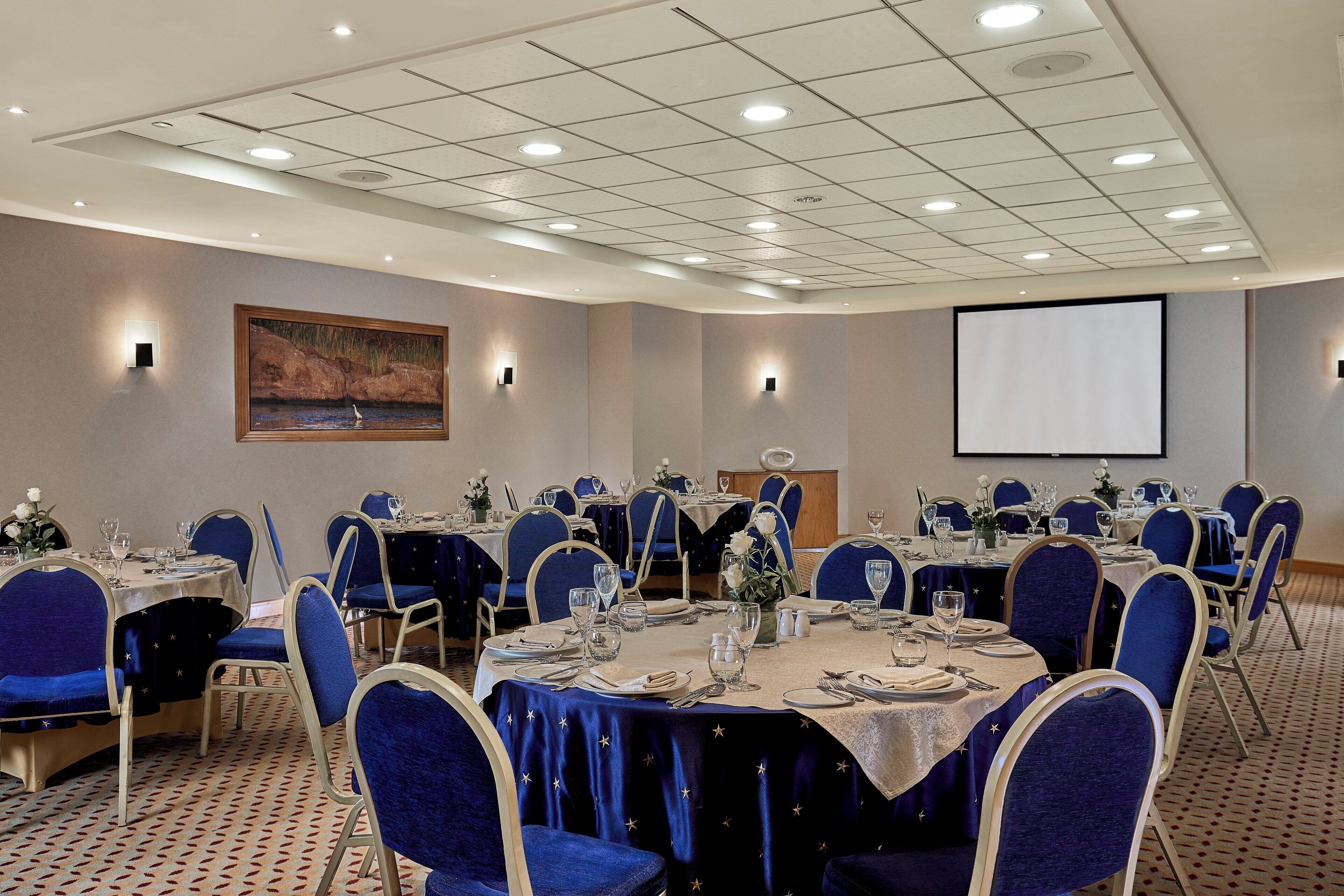 Un salón de conferencias con mesas redondas, sillas azules y una pantalla de proyector en la pared.