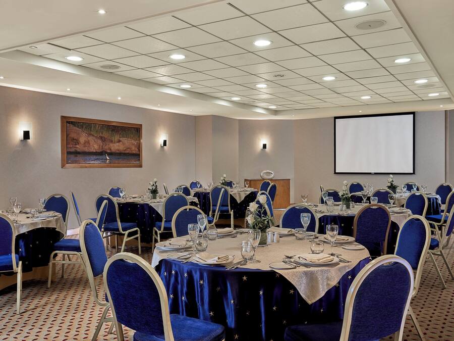 Un salón de conferencias con mesas redondas, sillas azules y una pantalla de proyector en la pared.