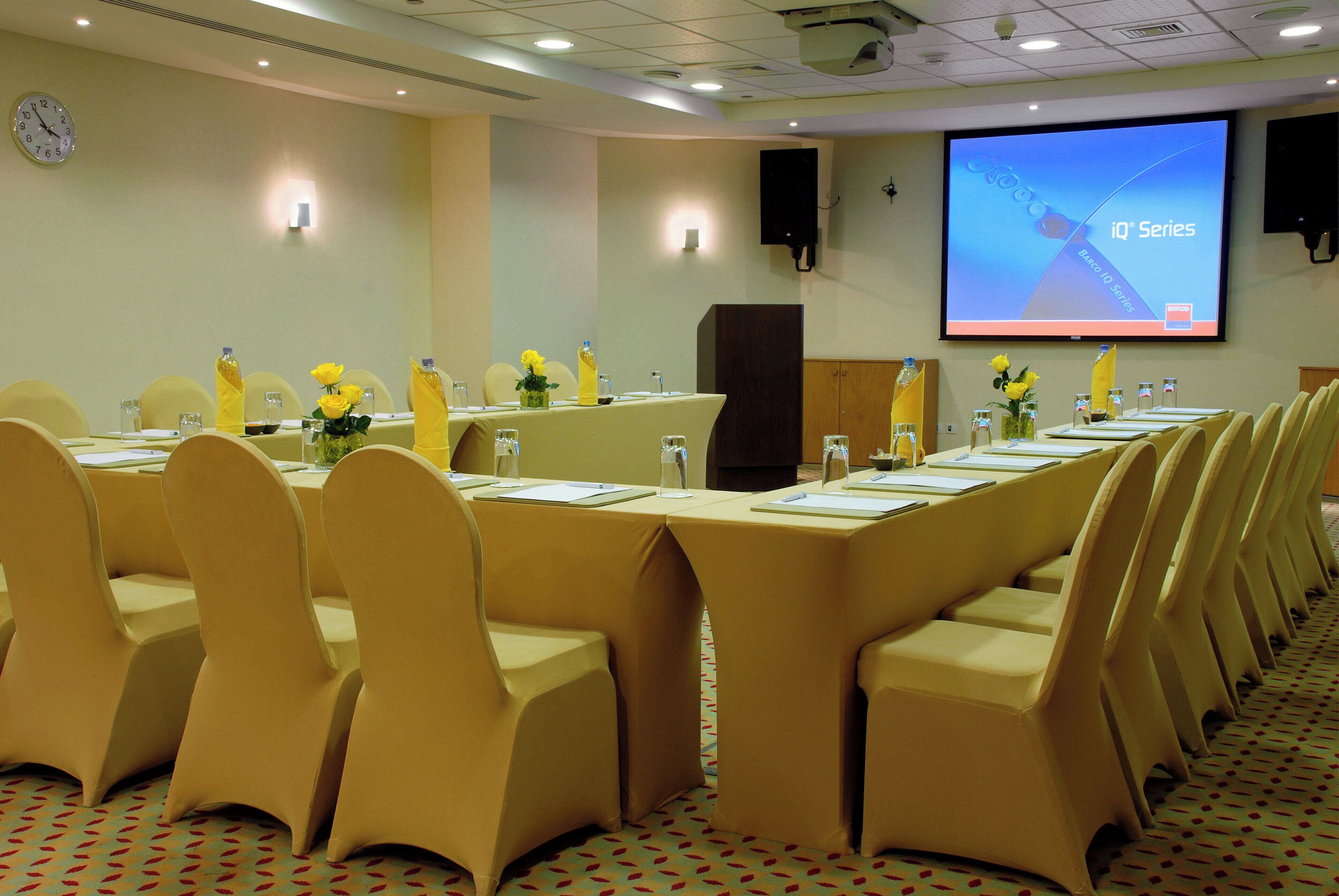 A conference room with a long table, chairs, a projector screen, and a clock on the wall.