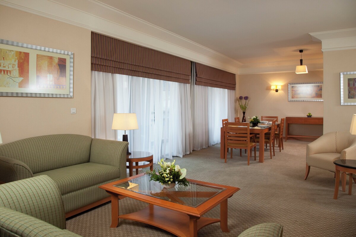 Une chambre d’hôtel avec un canapé vert, une table basse en verre, une table à manger avec des chaises, une lampe et un tableau au mur.