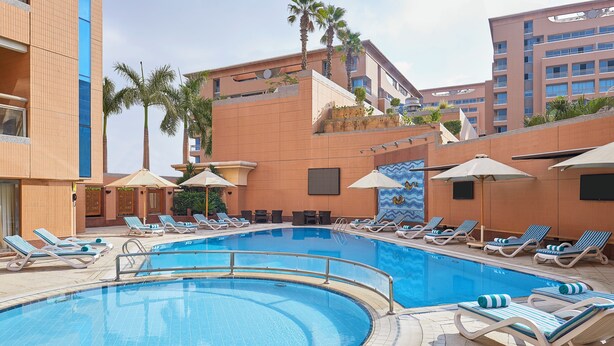 A pool area with lounge chairs and umbrellas, surrounded by buildings with balconies and palm trees. - Holiday Inn Cairo Citystars by IHG (Cairo)