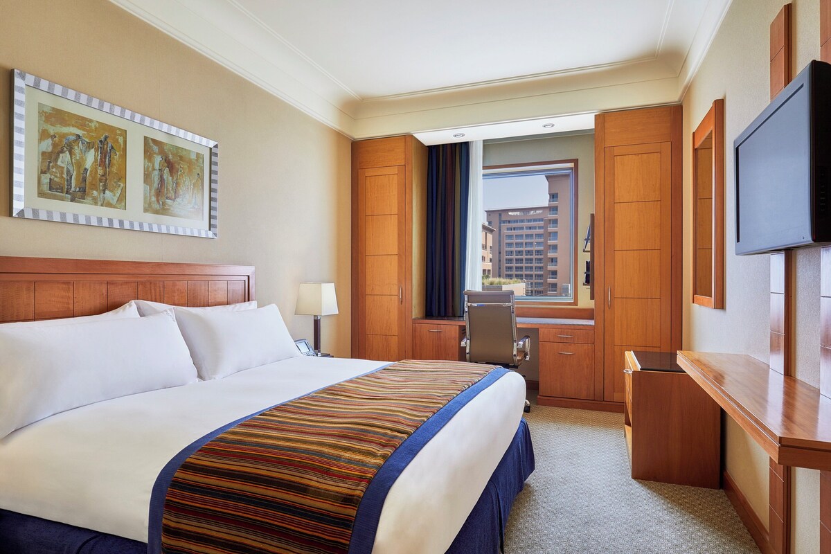 Une chambre d’hôtel avec un grand lit, un bureau, une chaise, une télévision et une fenêtre donnant sur des immeubles.