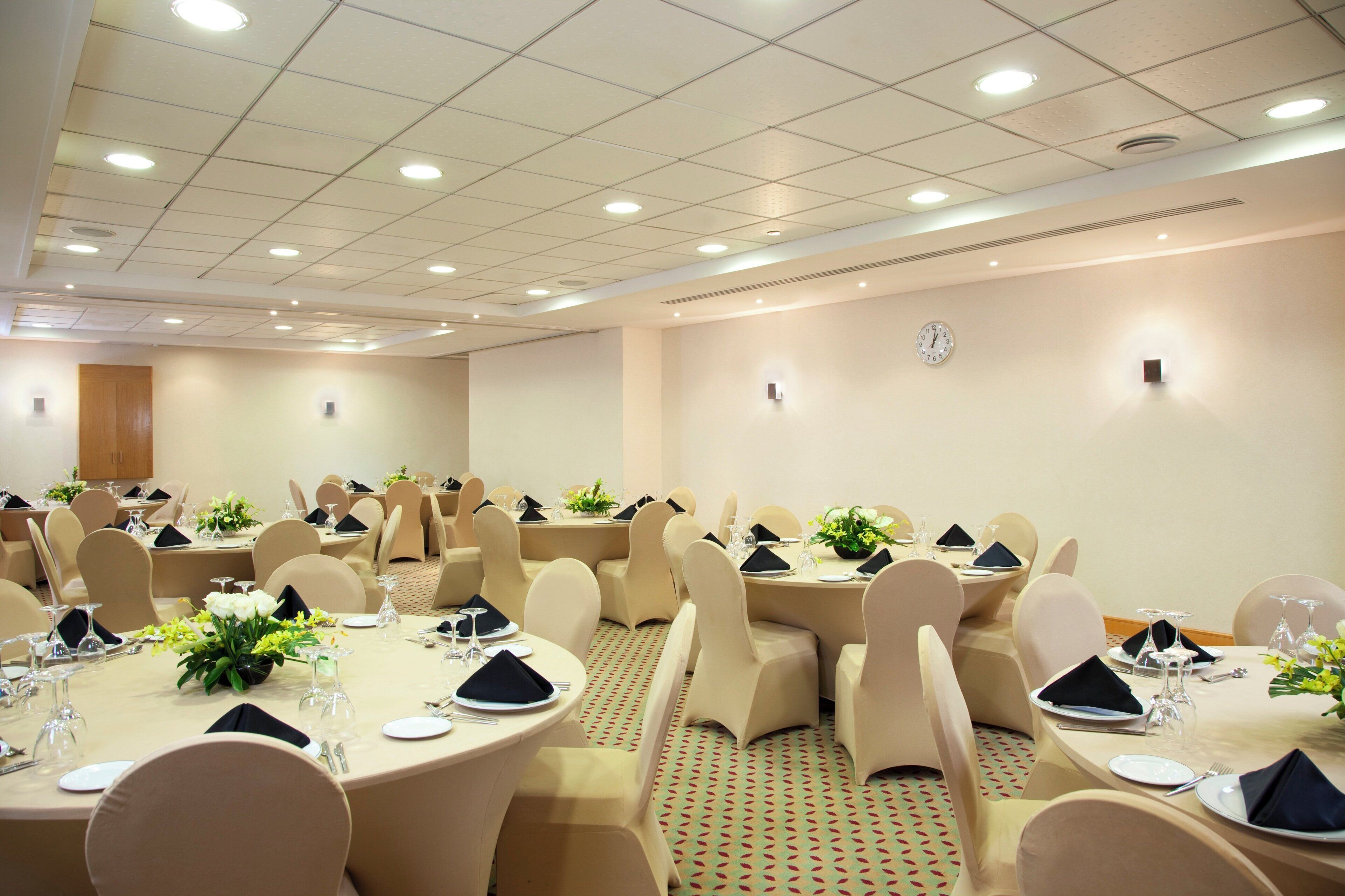 Een vergaderzaal met ronde tafels, beige stoelen en een klok aan de muur.