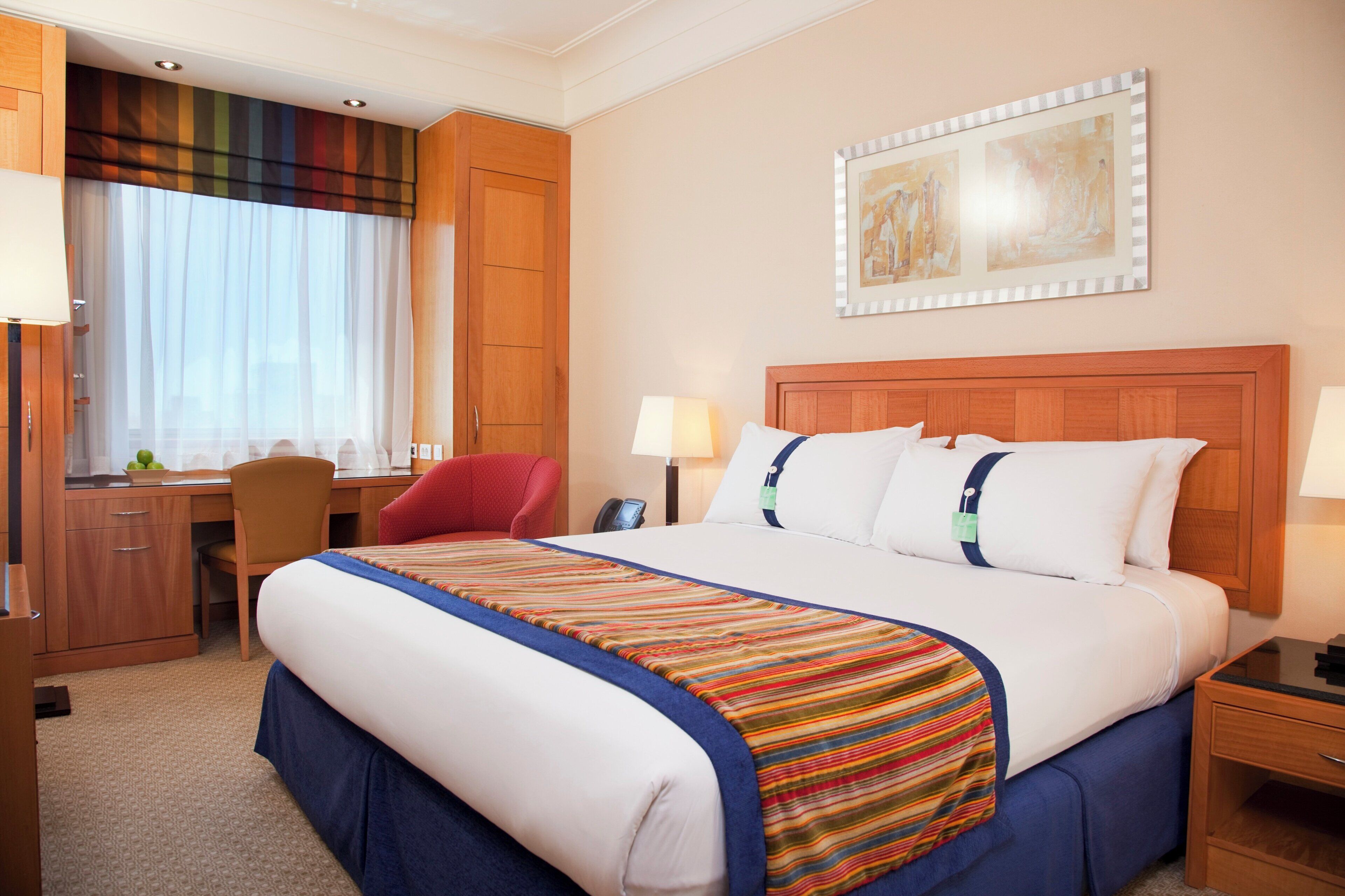 A hotel room with a bed, a desk, a chair, a nightstand, and a painting on the wall.