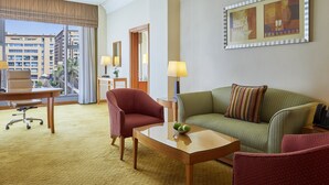 Une chambre d’hôtel avec un canapé vert, un fauteuil rouge, une table basse en bois avec des pommes, un bureau avec une lampe et une grande fenêtre donnant sur des immeubles.