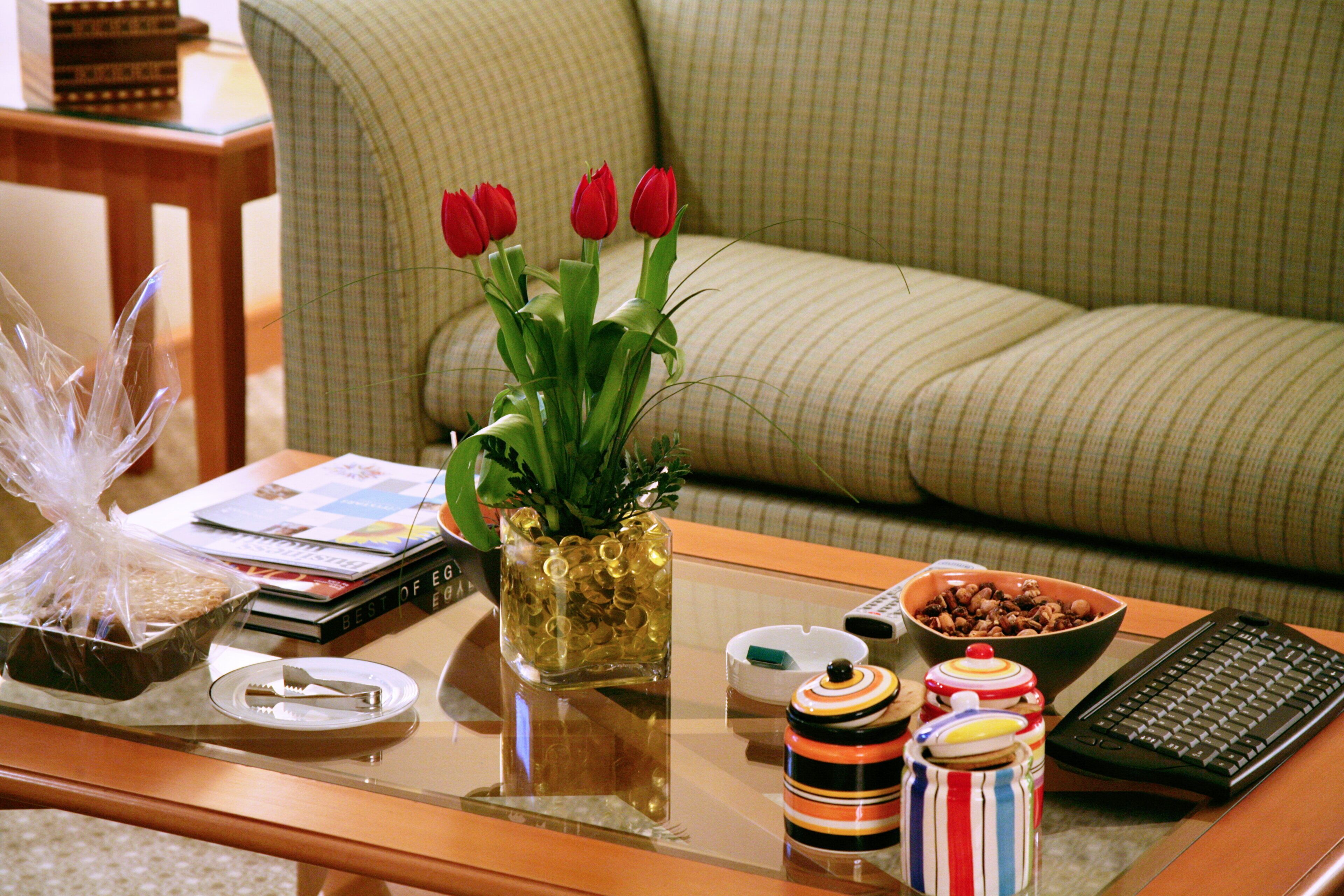 Una sala de estar con una mesa de centro de vidrio, un cuenco de nueces, una taza a rayas, un teclado y un jarrón con flores rojas.