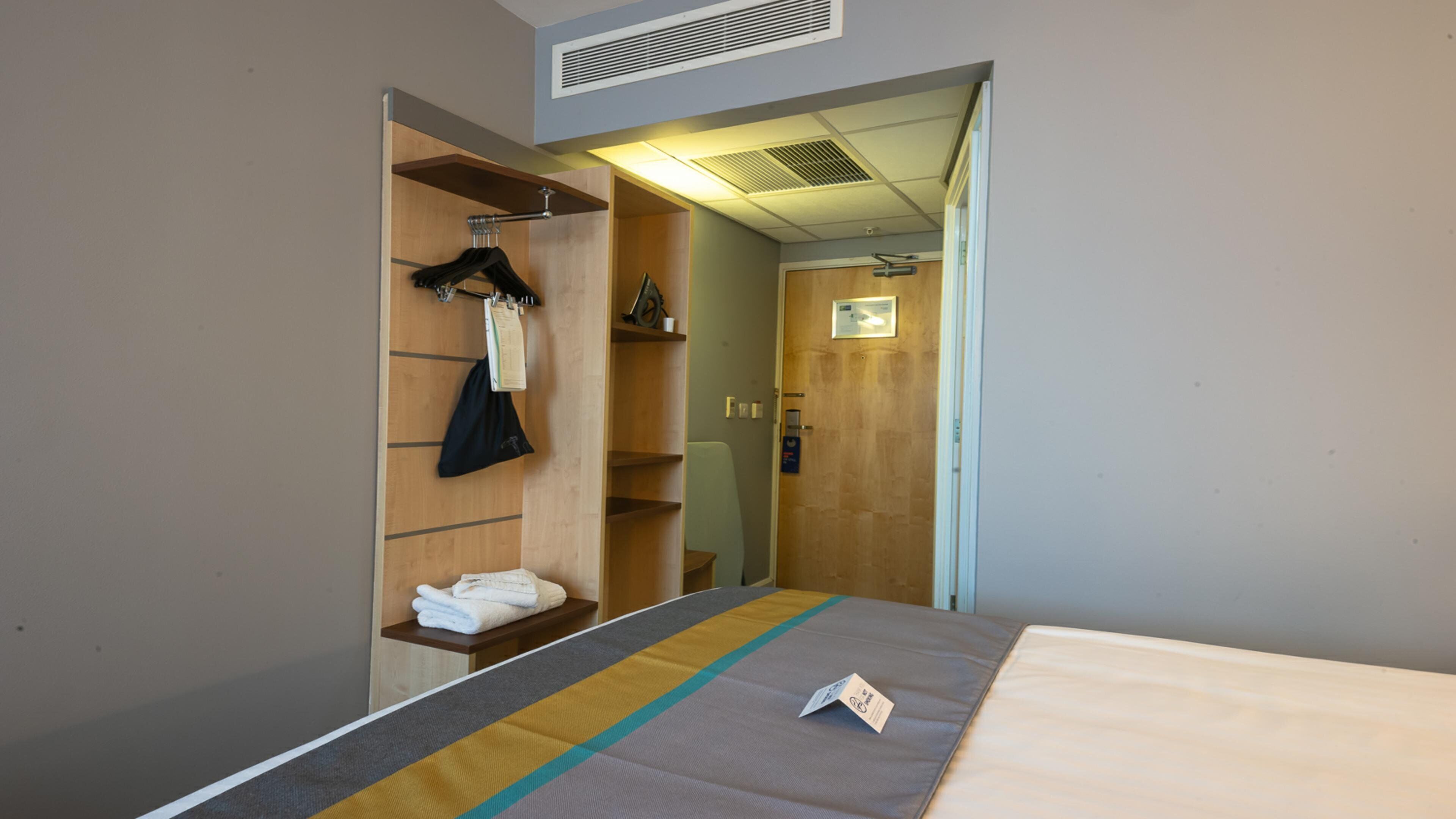 Habitación de hotel con cama, armario de madera, estante con toallas dobladas y puerta que da a otra habitación.