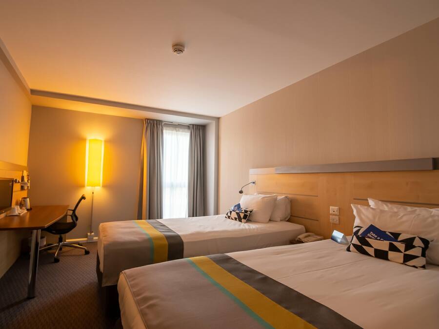 A hotel room with two beds, a desk, a chair, a lamp, and a window with curtains.
