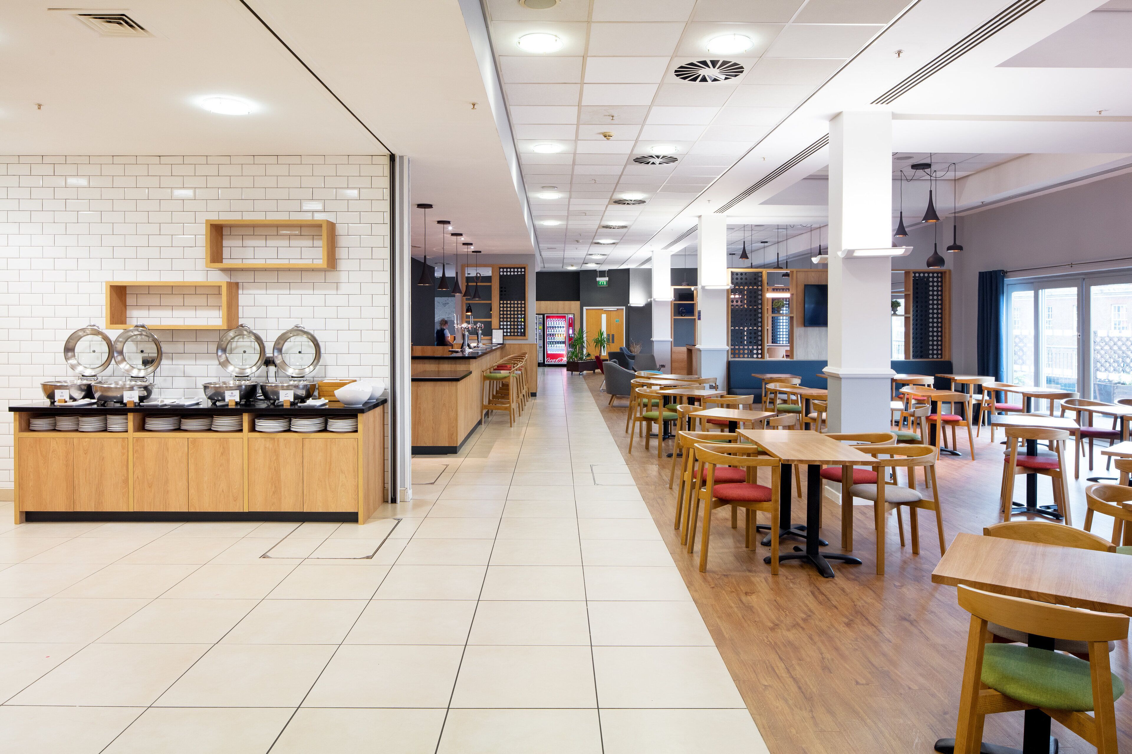 A spacious cafe with wooden tables and chairs, a counter with a coffee machine, and a tiled floor.