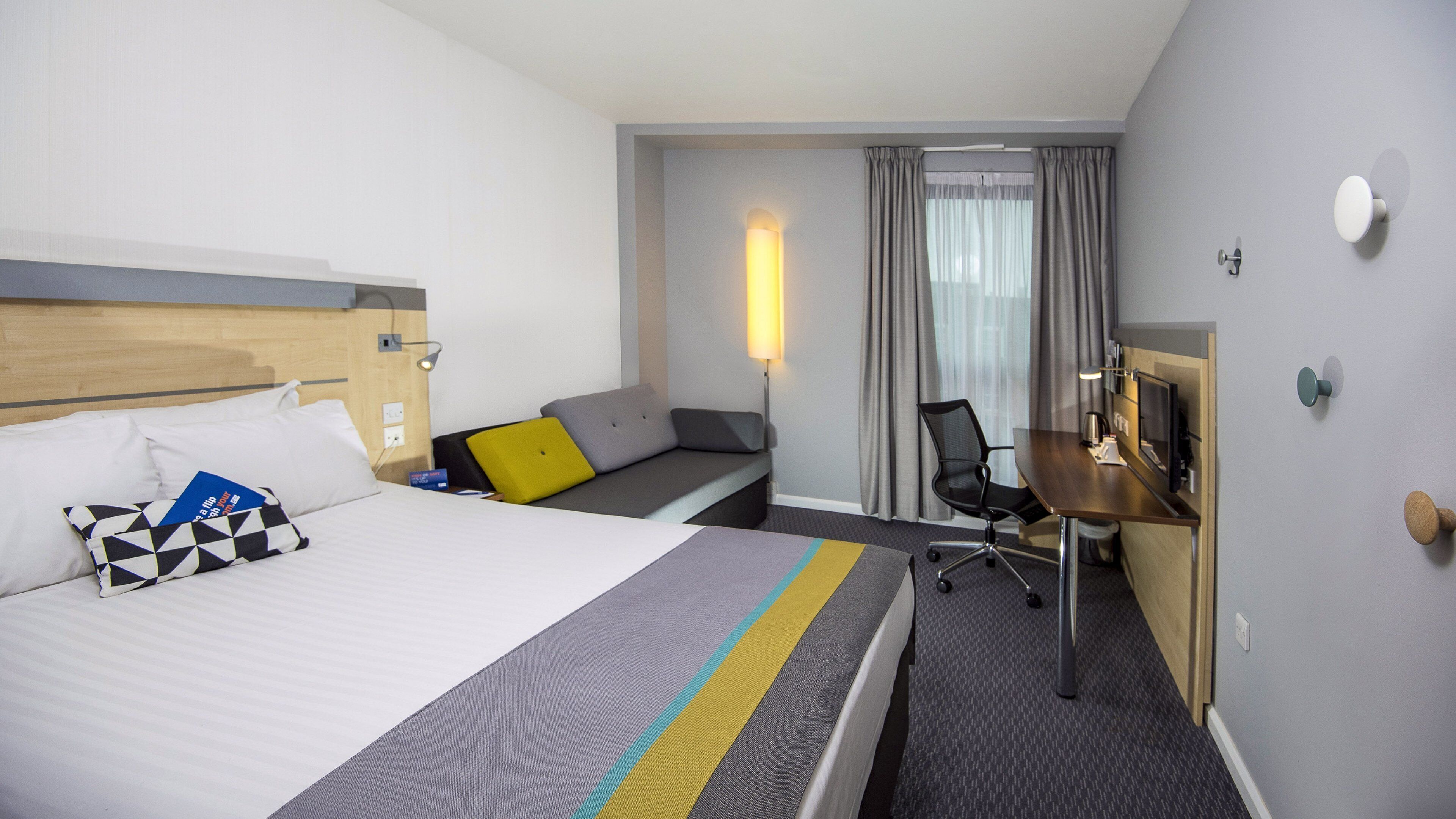 Una habitación de hotel con cama, escritorio, silla y ventana con cortinas.