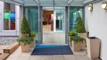 Entrada de hotel con puerta de vidrio, felpudo azul y plantas en macetas.