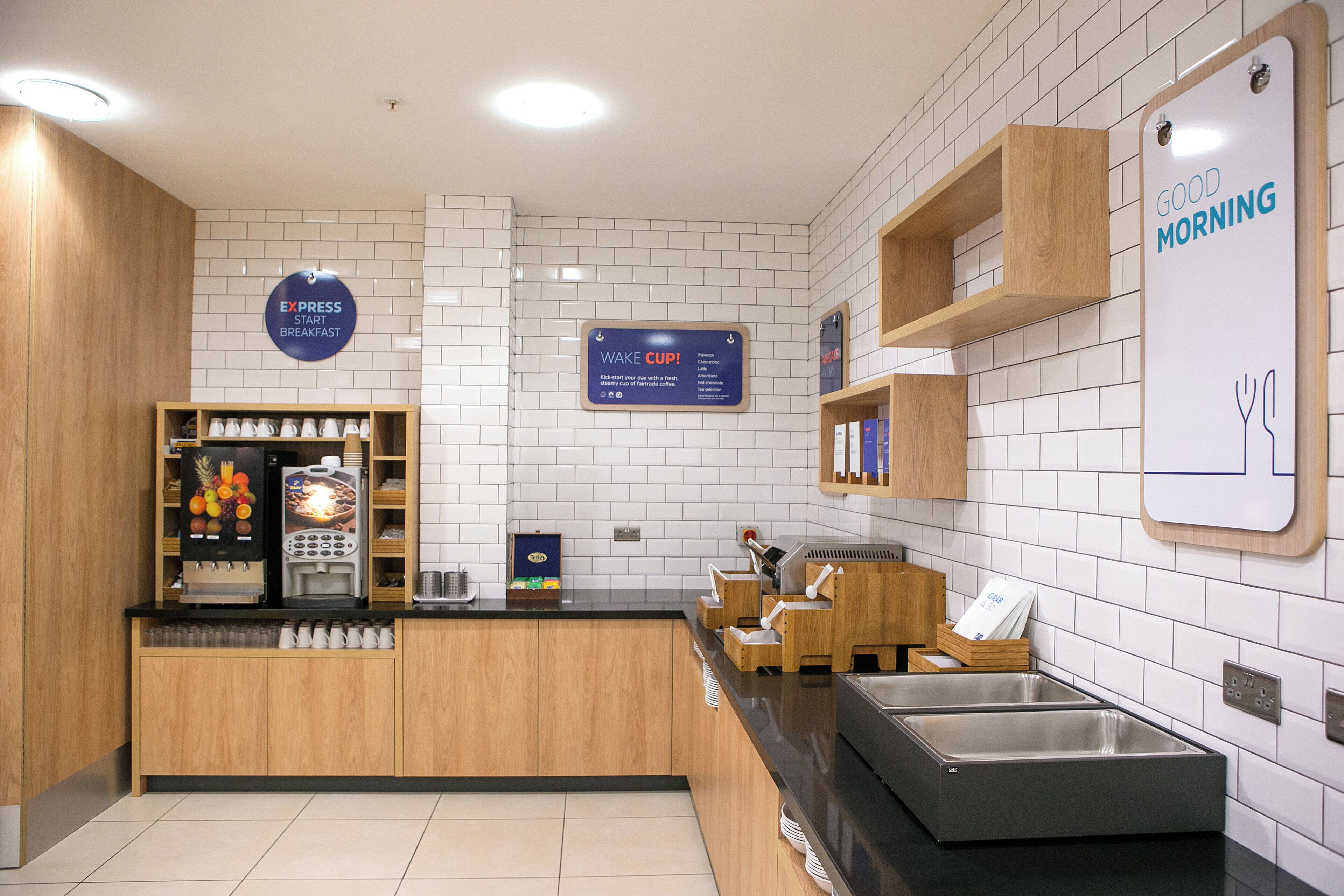 A modern breakfast area with a coffee machine, a fruit display, and a sign indicating breakfast options.