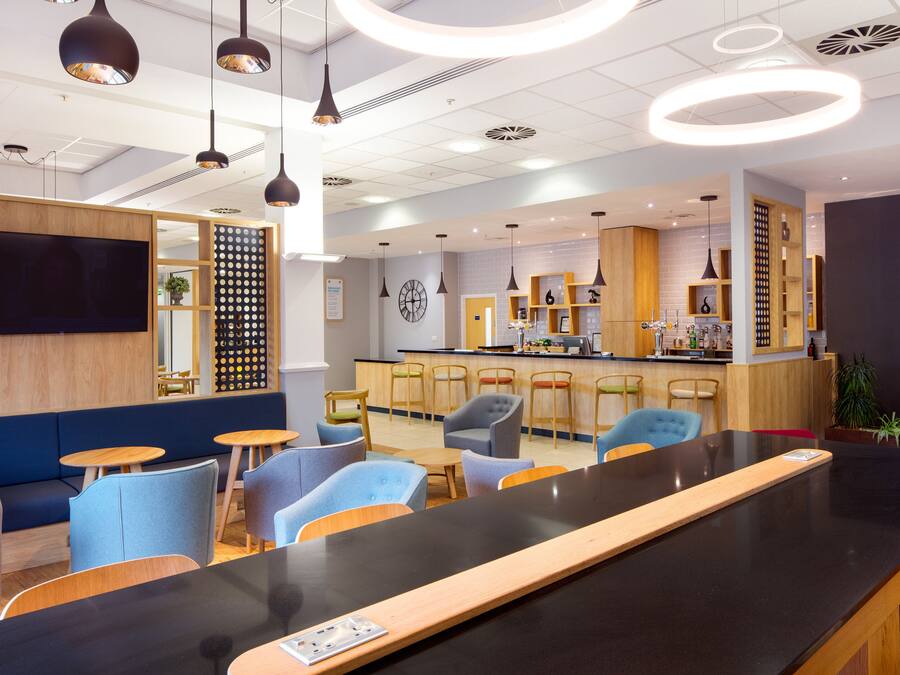 Een moderne bar met een lange toonbank, verschillende zitplaatsen zoals barstoelen en gewone stoelen, en een zichtbare keuken op de achtergrond.