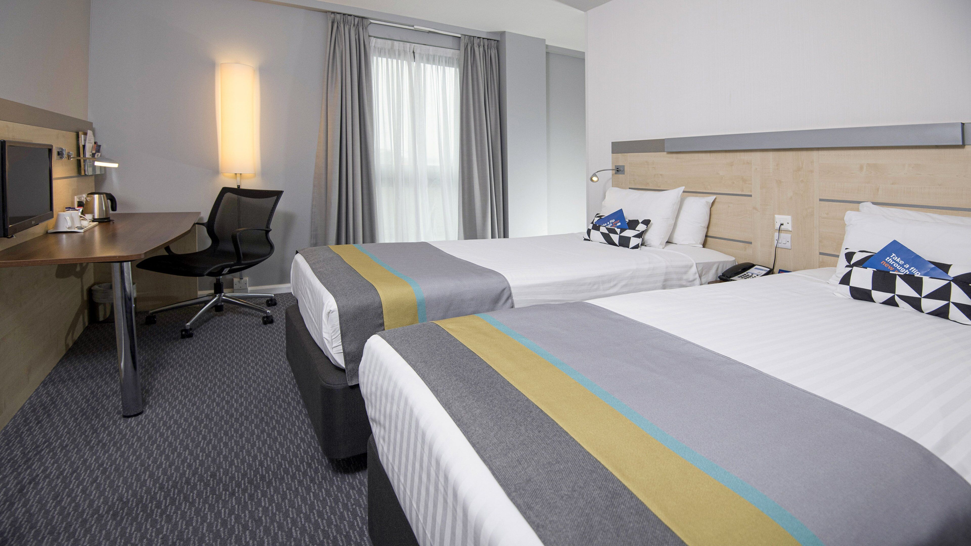 A hotel room with two beds, a desk, a chair, a television, and a window with curtains.
