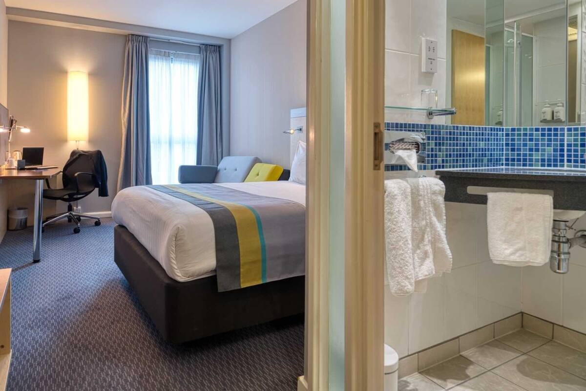 Une chambre d’hôtel comprenant un lit, un bureau avec une chaise, une lampe et une salle de bain avec un miroir et un lavabo.