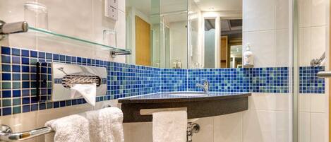 A bathroom with a blue-tiled wall, a sink with a mirror above it, and a glass shelf with toiletries.