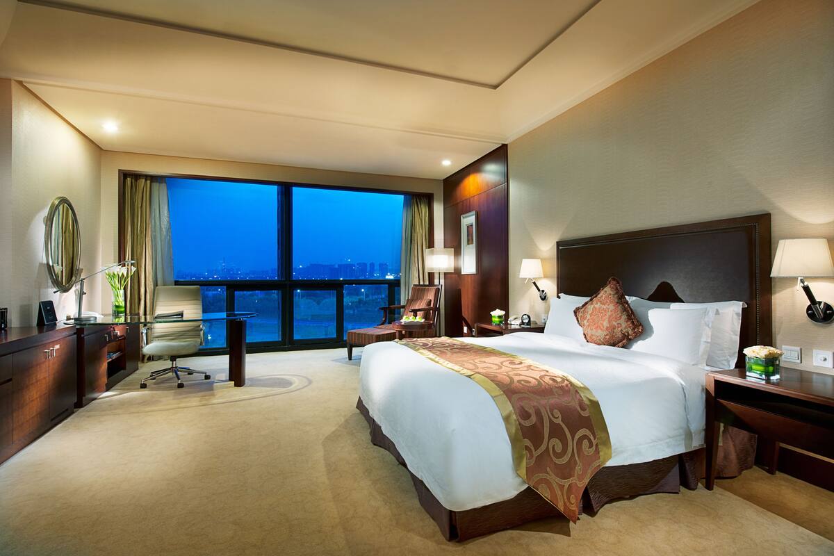 Habitación de hotel con una cama grande, un escritorio, una silla y vistas a la ciudad al anochecer.