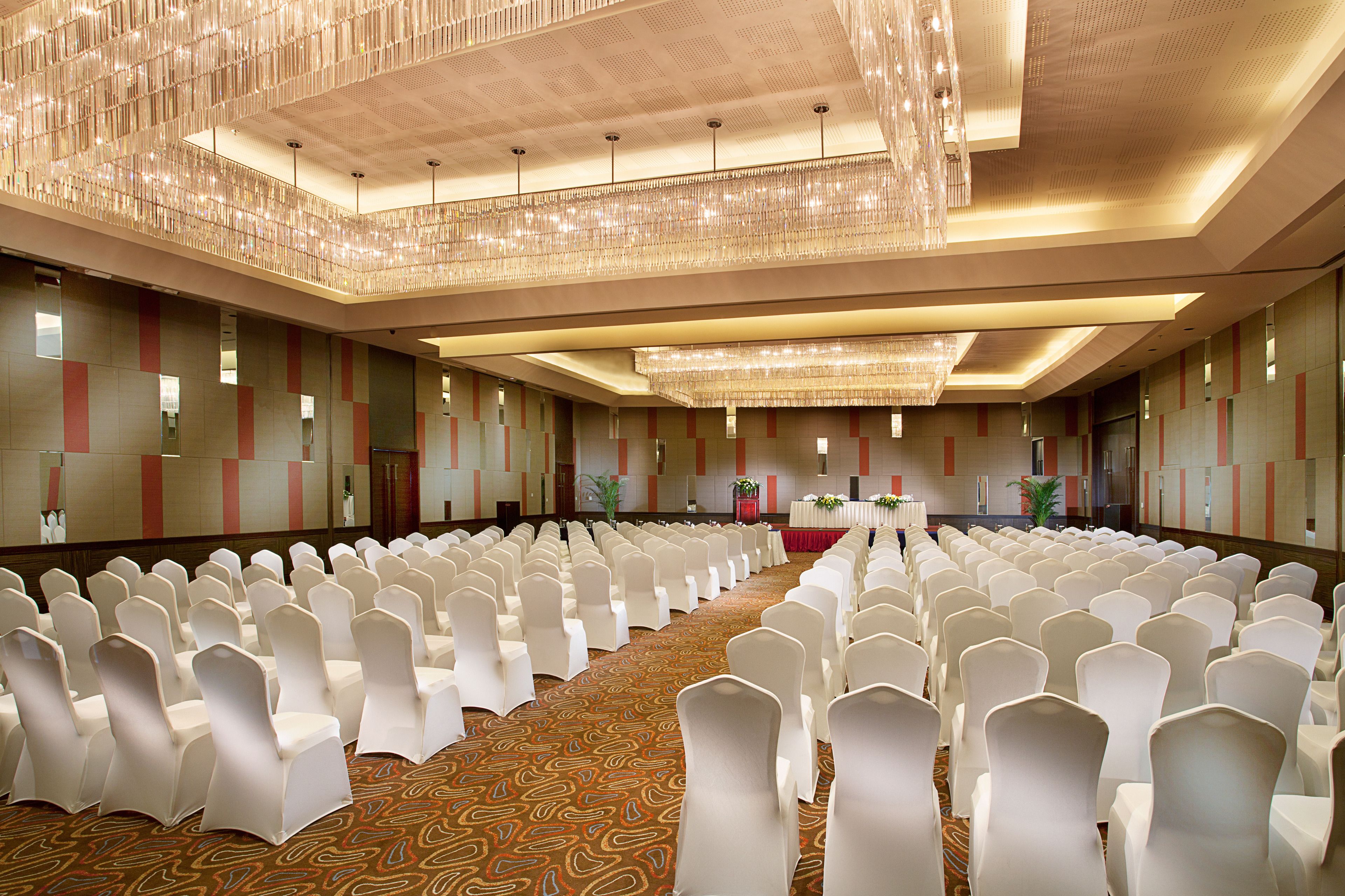 A large conference room with rows of white chairs, a stage with a red table, and a chandelier.