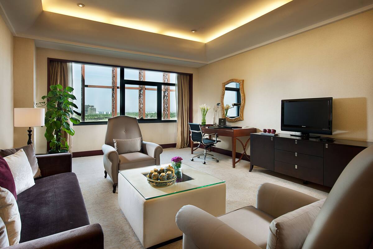 Habitación de hotel con sofá, sillones, una mesa de centro, televisor, escritorio y un amplio ventanal con vistas a la ciudad.