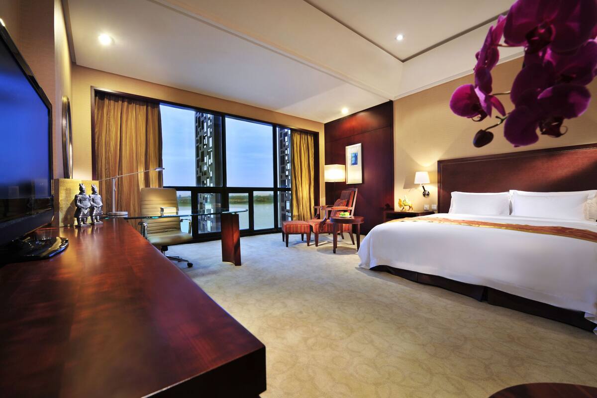 Habitación de hotel con una cama grande, un escritorio con computadora, una silla y un ventanal con cortinas.