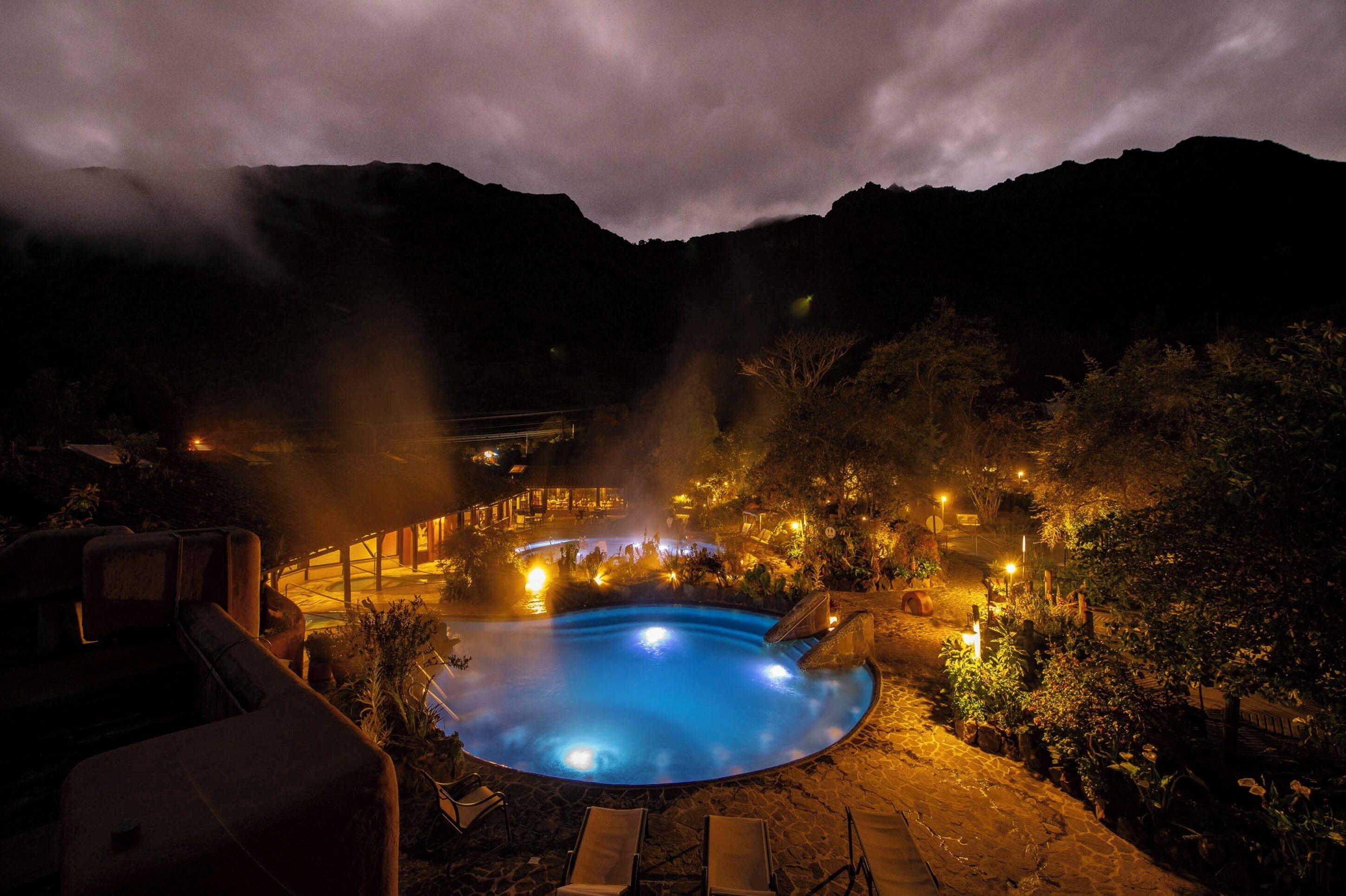 A hot spring surrounded by a wooden deck and stone pathway, with a mountainous backdrop.