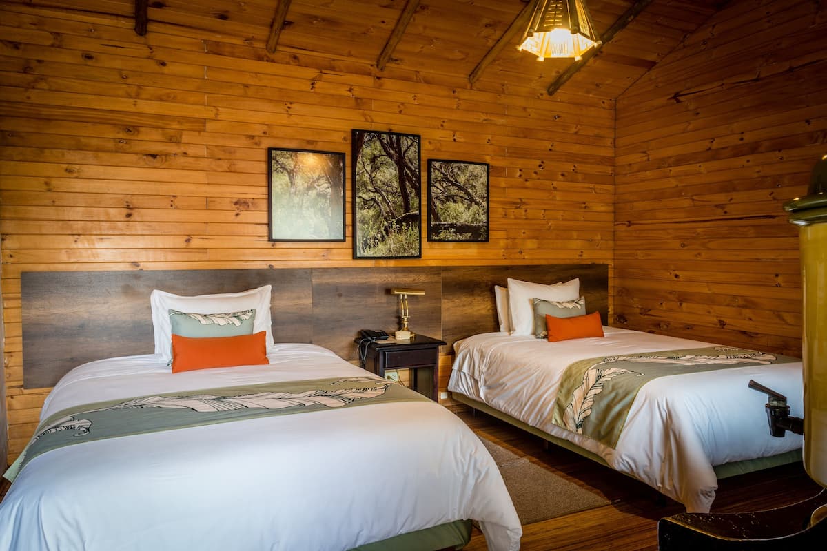 A wooden cabin room with two beds, a nightstand, a lamp, and framed pictures on the wall.