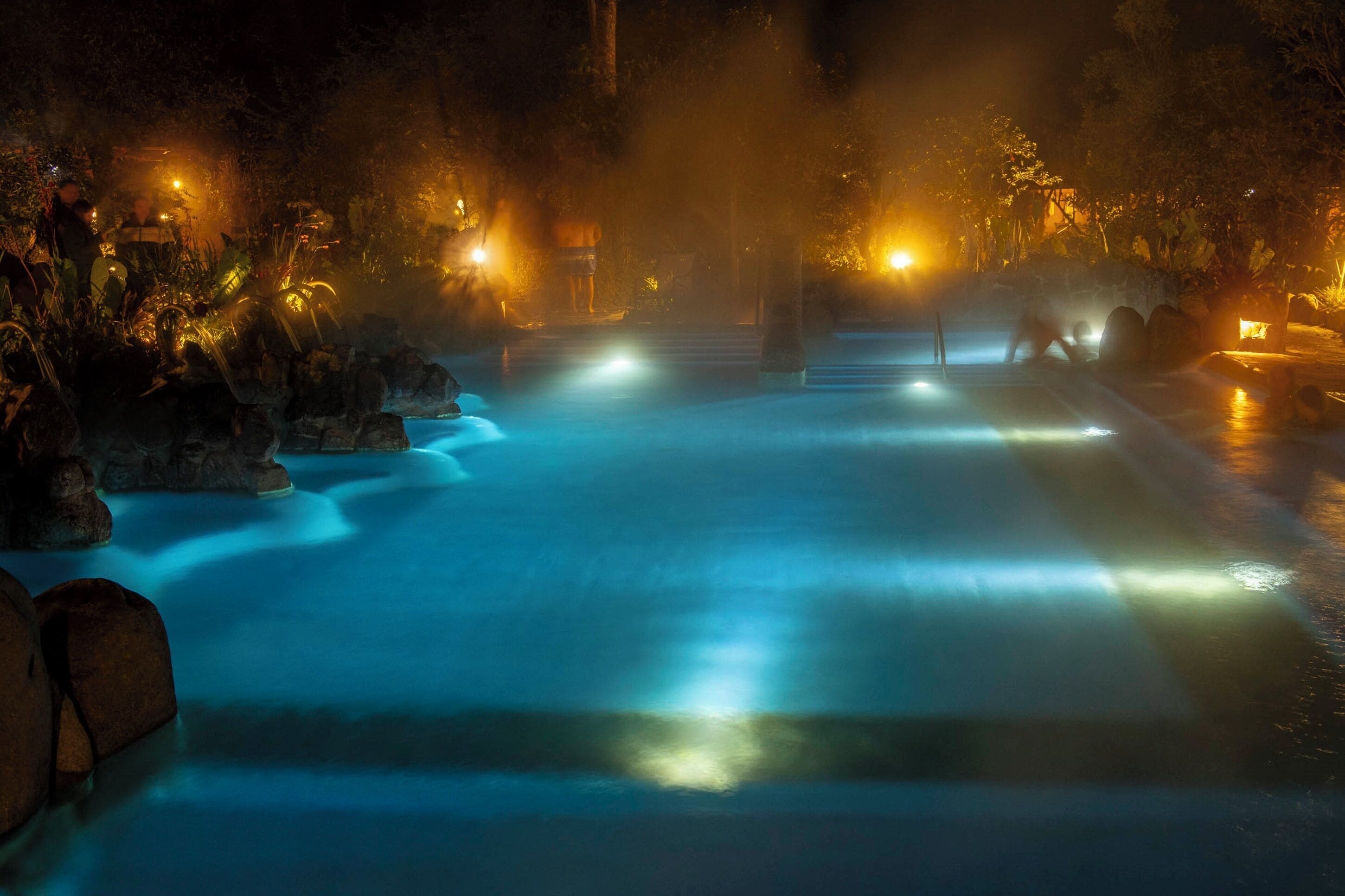 Un área de aguas termales nocturna con senderos iluminados y vegetación circundante.