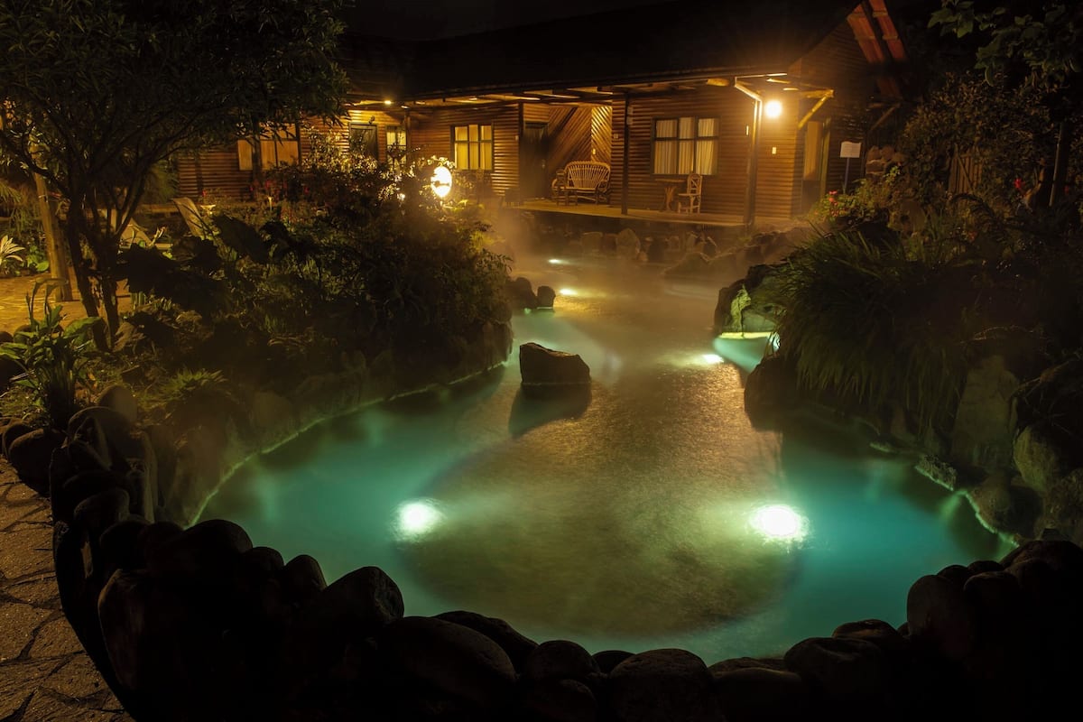 A nighttime view of a hot spring area with a wooden cabin and a stone pathway leading to the water.
