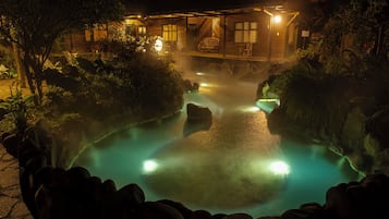 Vista nocturna de una zona de aguas termales con una cabaña de madera y un sendero de piedra que conduce al agua.