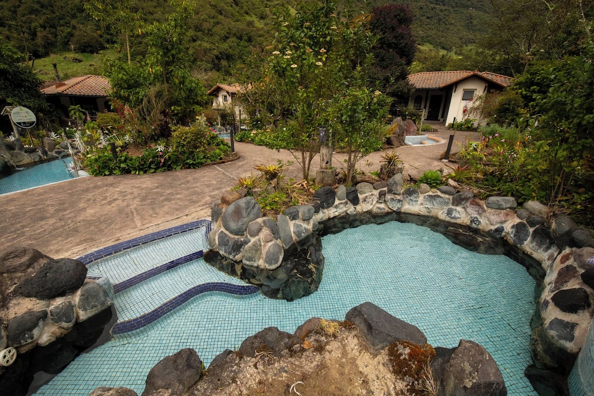 A pool with a natural rock border, surrounded by lush greenery and a small building with a tiled roof.
