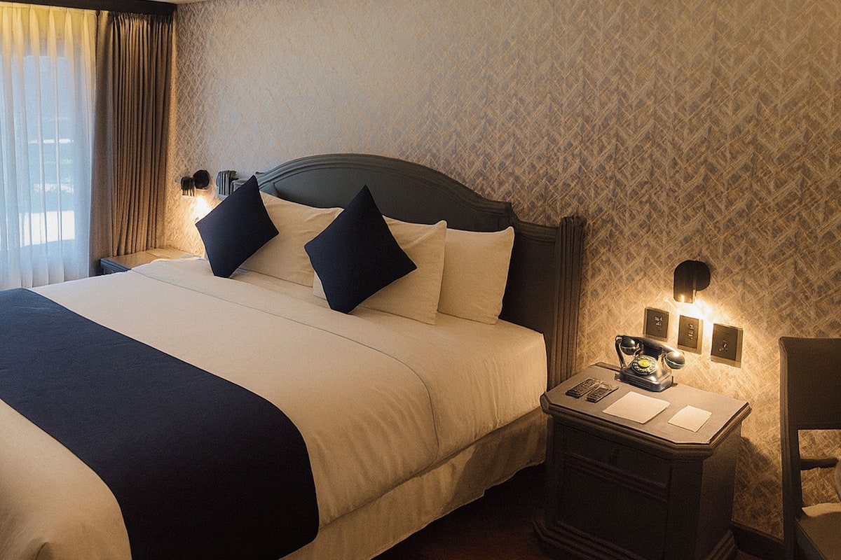 A neatly made bed with a dark headboard, a bedside table with a lamp, and a patterned wallpaper.